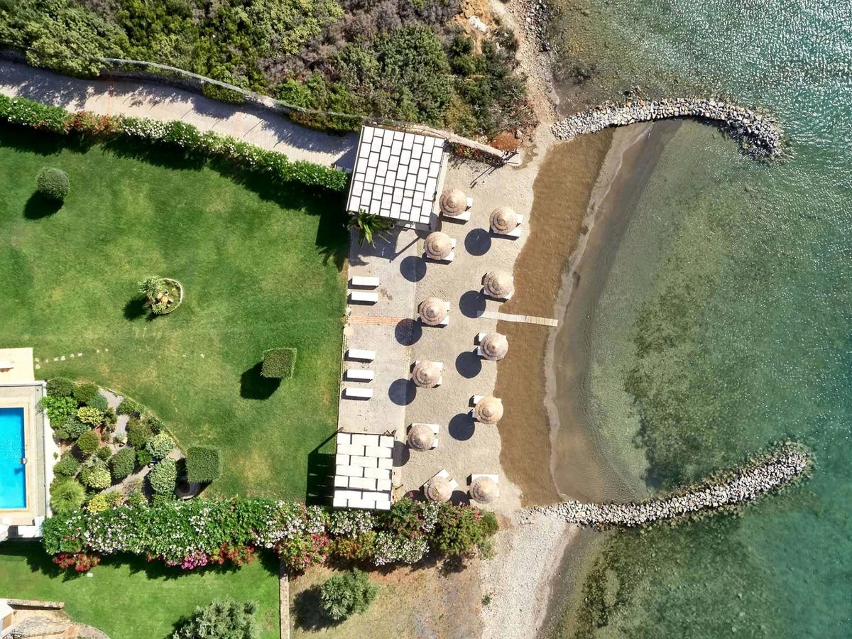 Aerial view straight down onto the beach club: thatched umbrellas and loungers on a strip of sand between two stone breakwaters enclosing a shallow cove.