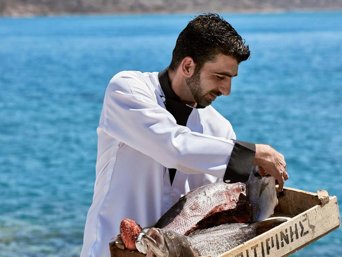 A chef in a white jacket holding a wooden crate of whole fresh fish in front of the blue sea.