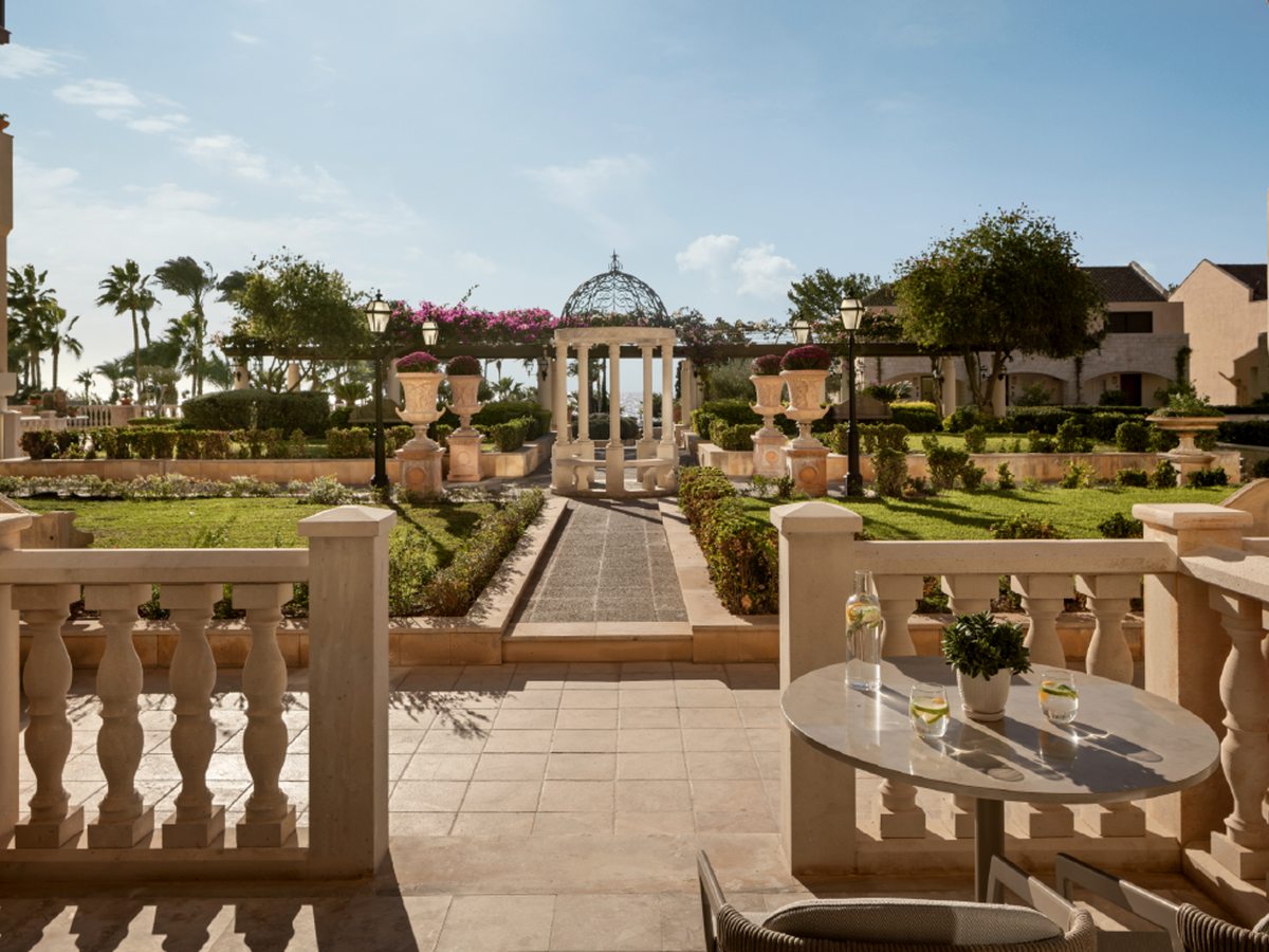 A formal garden with clipped hedges, a domed stone gazebo and flower-filled urns, seen from a shaded terrace.