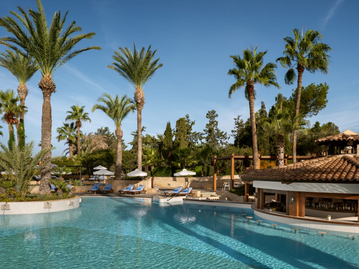 A large freshwater pool ringed by tall palm trees, with a thatched swim-up bar on the right and sunloungers under parasols.