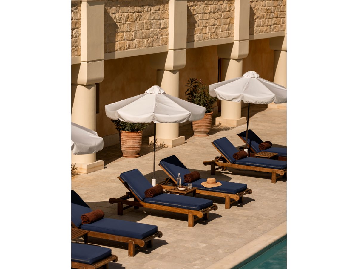 Wooden sunloungers with navy cushions and white parasols on a stone terrace beside the pool, below a honey-stone colonnade.