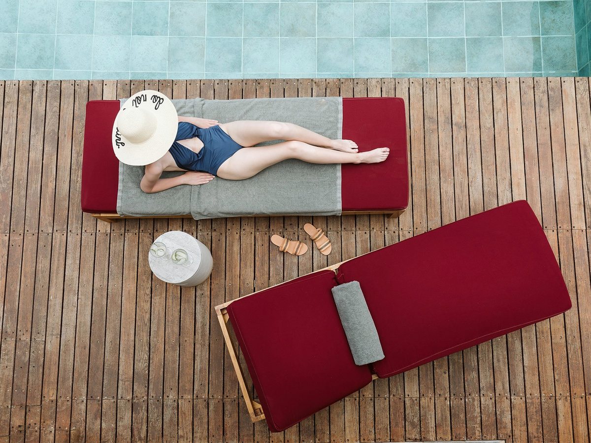 An overhead view of a timber sundeck at the pool's edge, a red-cushioned lounger, a sun hat and a small side table.