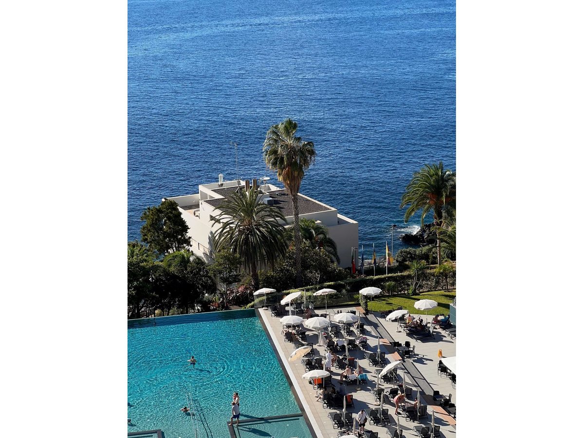 The pool terrace in daylight, rows of sun loungers under white parasols along the outdoor pool above the sea, with palms and the Atlantic beyond.