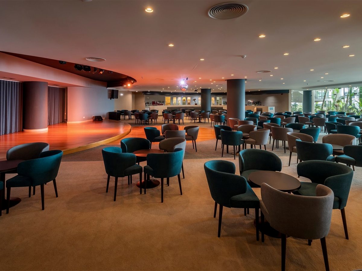 A large hotel lounge with a wooden dance floor and a curved stage area, a bar at the back and clusters of teal and grey tub chairs around low round tables on a beige carpet.