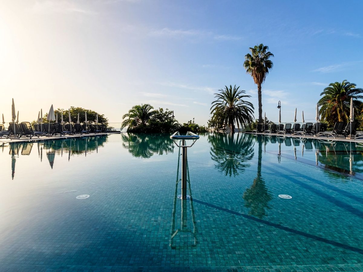 The outdoor infinity pool on a sunny morning, its still water reflecting palm trees, with sun loungers and parasols along both sides and a metal shower post in the centre.