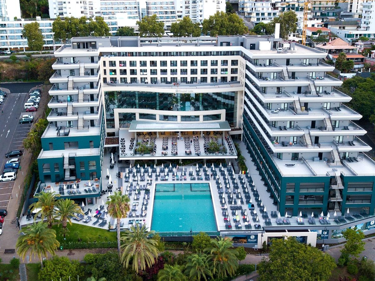Aerial view of the Enotel Lido building, a large white and dark-green hotel block, with a rectangular pool and rows of sun loungers on the terrace in front, palm gardens around it and the town rising behind.