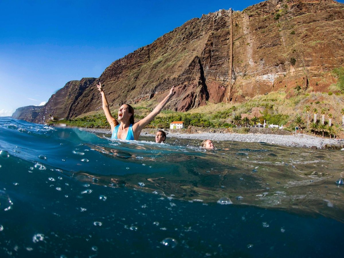 Three people swimming in clear blue sea just off a pebble shore, with tall terraced cliffs and banana plantations rising behind.