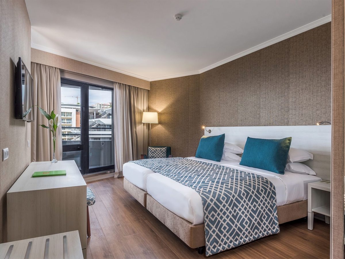 Guest room with a double bed dressed in white with a patterned throw and teal cushions, a wall-mounted TV, a desk and sliding doors onto a private balcony.