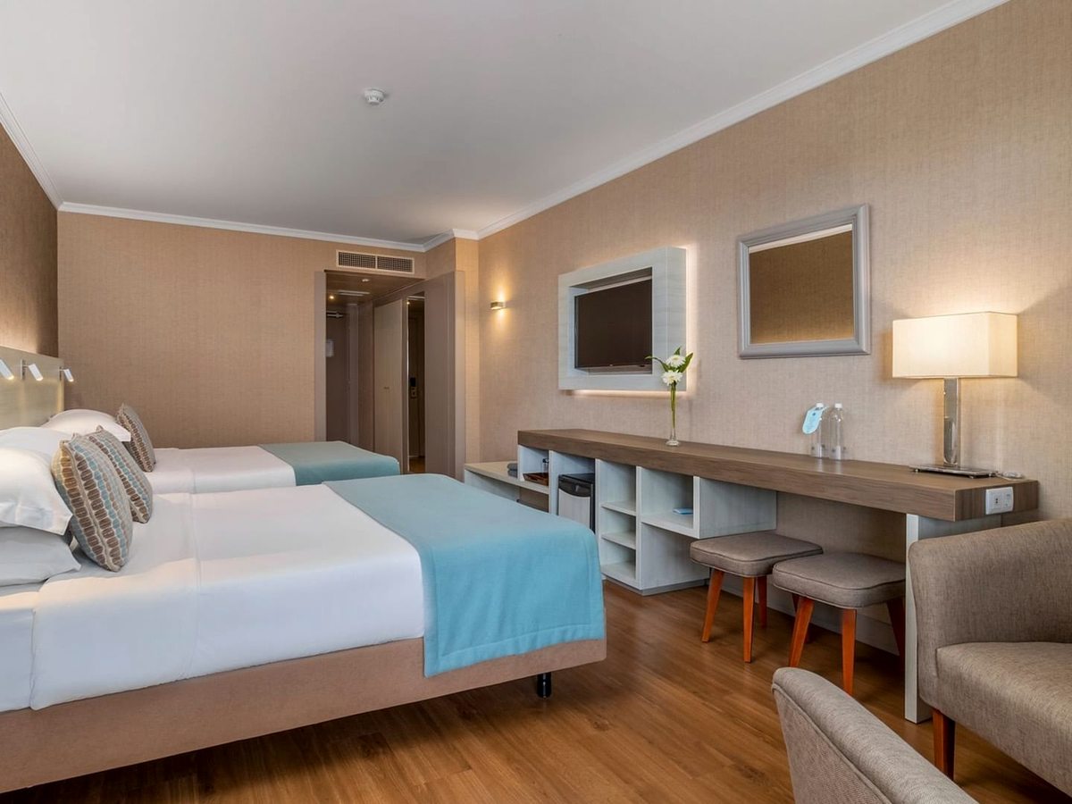Guest room with two single beds, a long desk and console with two stools, an armchair, a wall-mounted TV and a framed mirror, in warm beige tones.