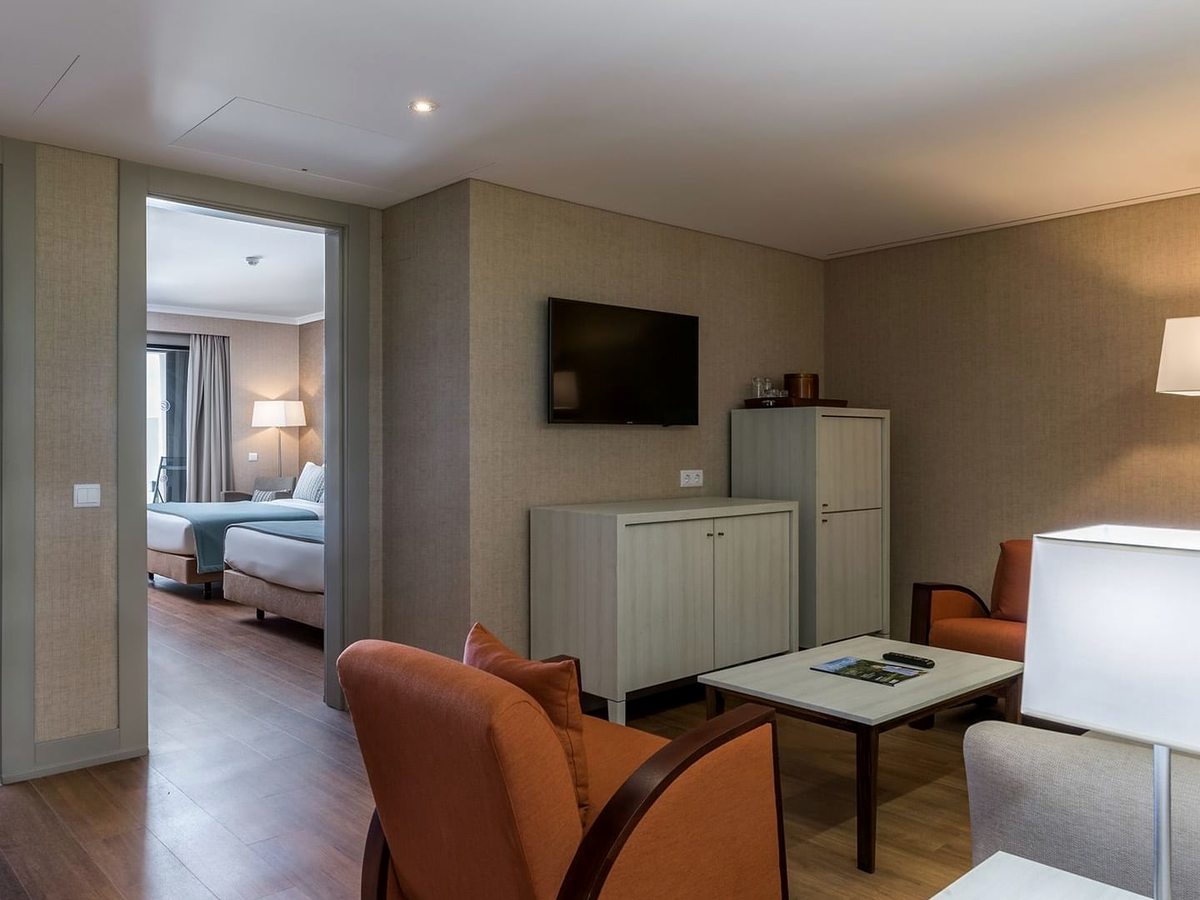 Suite living room with orange tub chairs, a coffee table, a wall-mounted TV and a sideboard, with the bedroom visible through an open doorway.