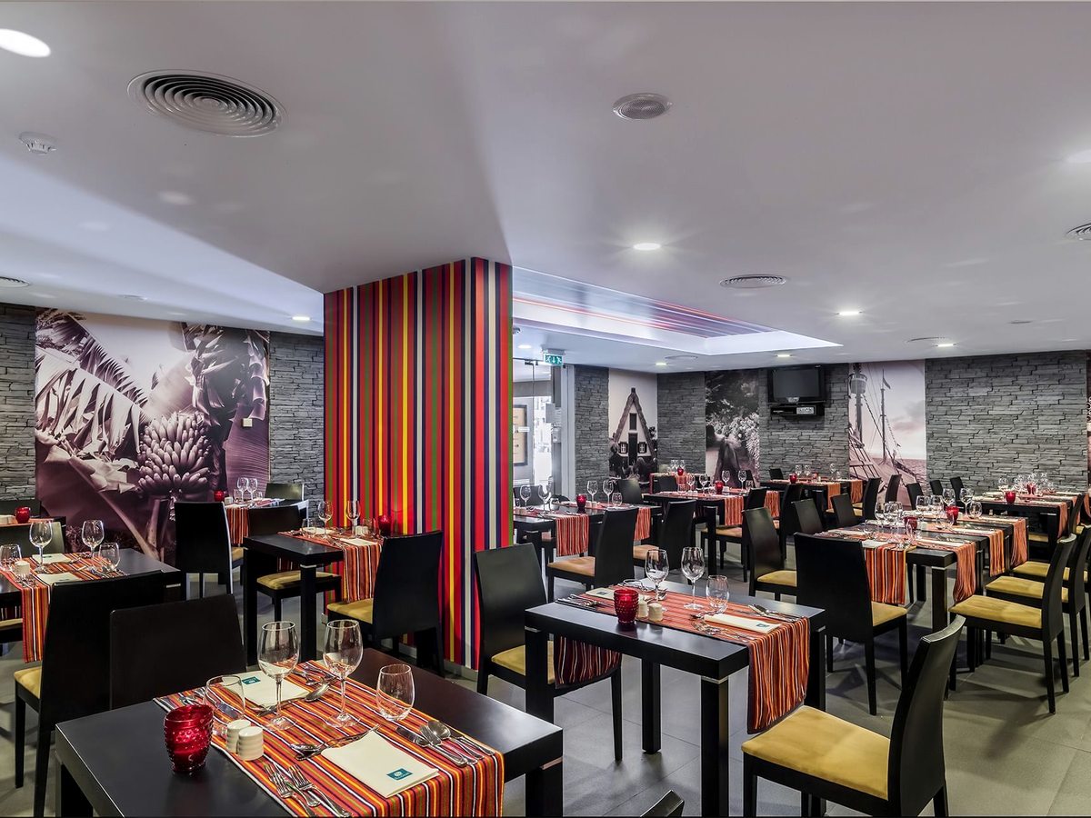 A restaurant dining room with black tables set with striped runners, wine glasses and place settings, and Madeiran-themed murals on the stone walls.