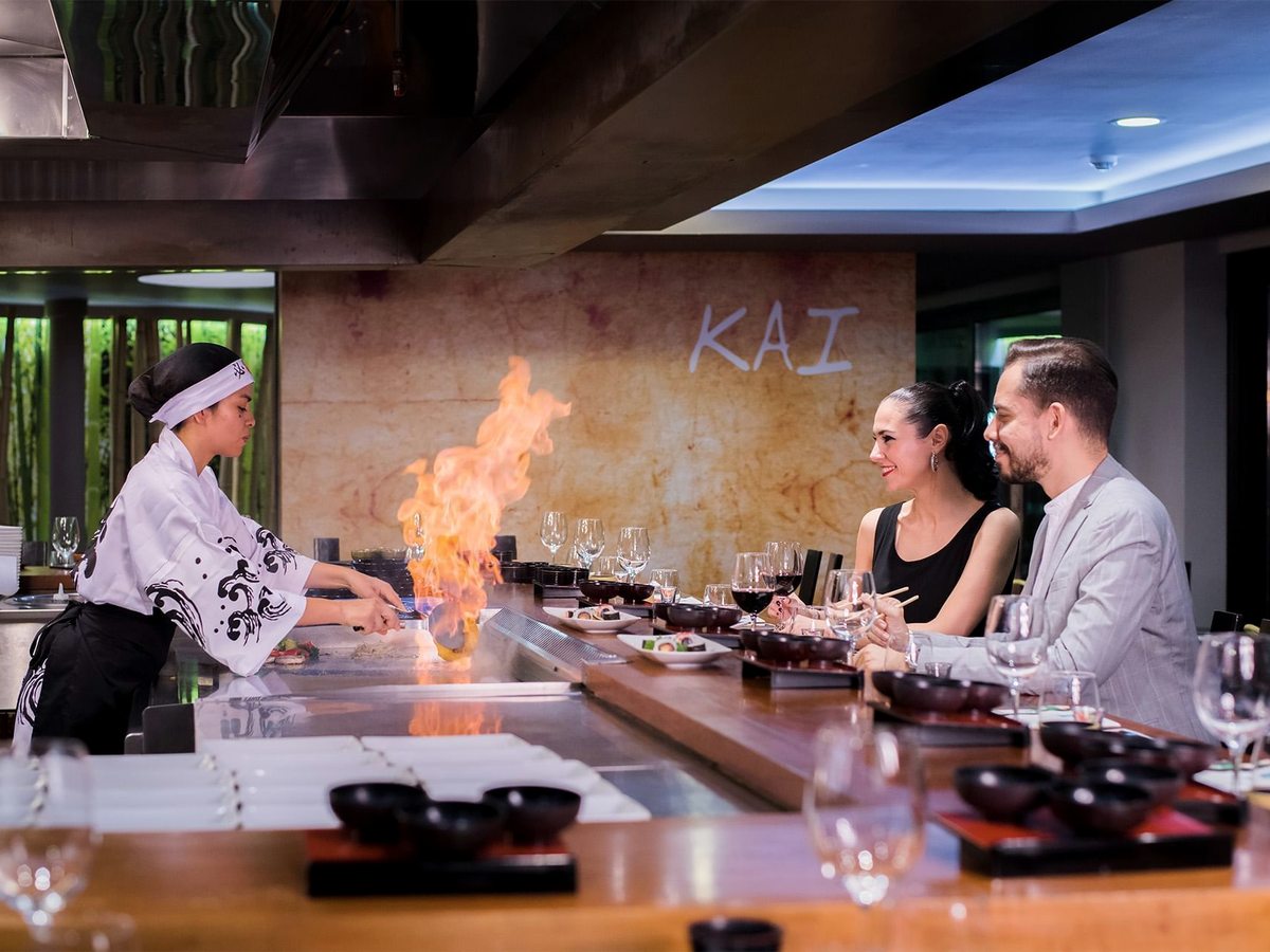 A chef flame-grilling at the teppanyaki counter at Kai, with two diners seated opposite and the word KAI on the wall behind.