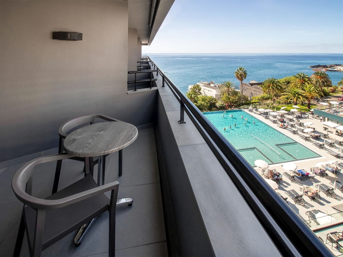 View from a room balcony with a small table and chair, looking down over the hotel's outdoor pool and lounger terrace and out to the Atlantic, with palms and the Funchal coast beyond.