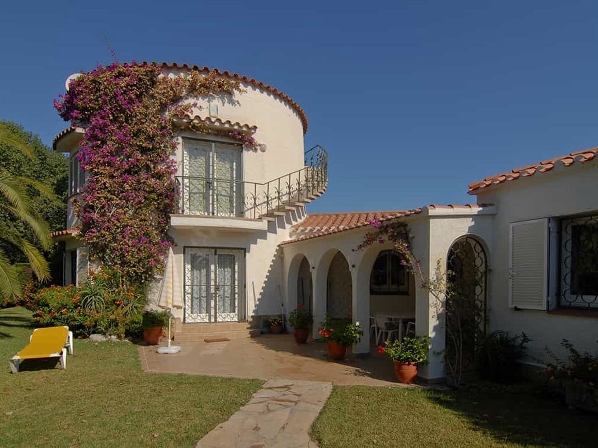 A white two-storey villa with a round bougainvillea-draped turret, an arched porch and a garden with a sun lounger under a clear blue sky.