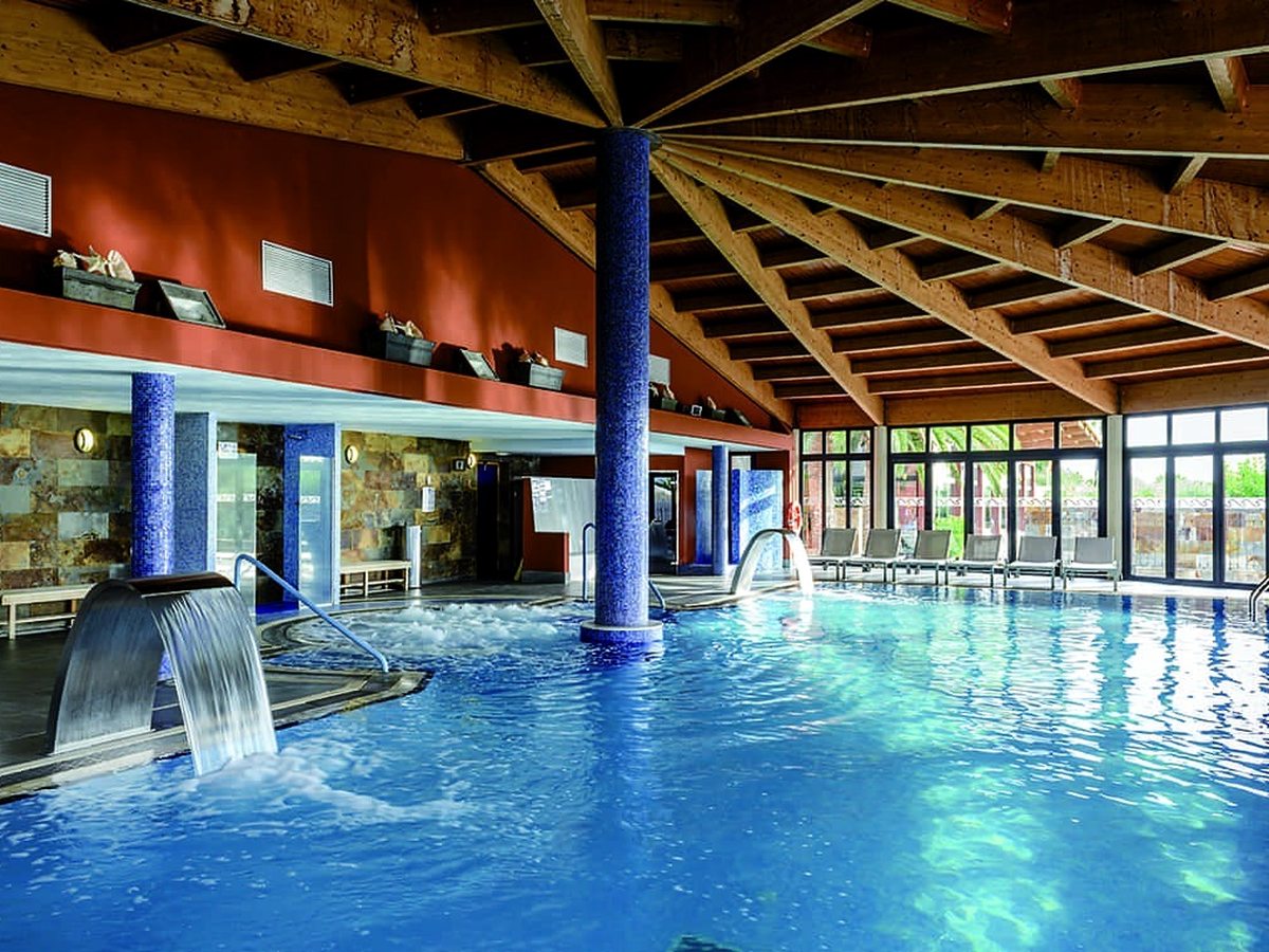 A heated indoor pool with a water-spout feature, blue mosaic columns and a high timber-beamed ceiling, with loungers by tall windows.