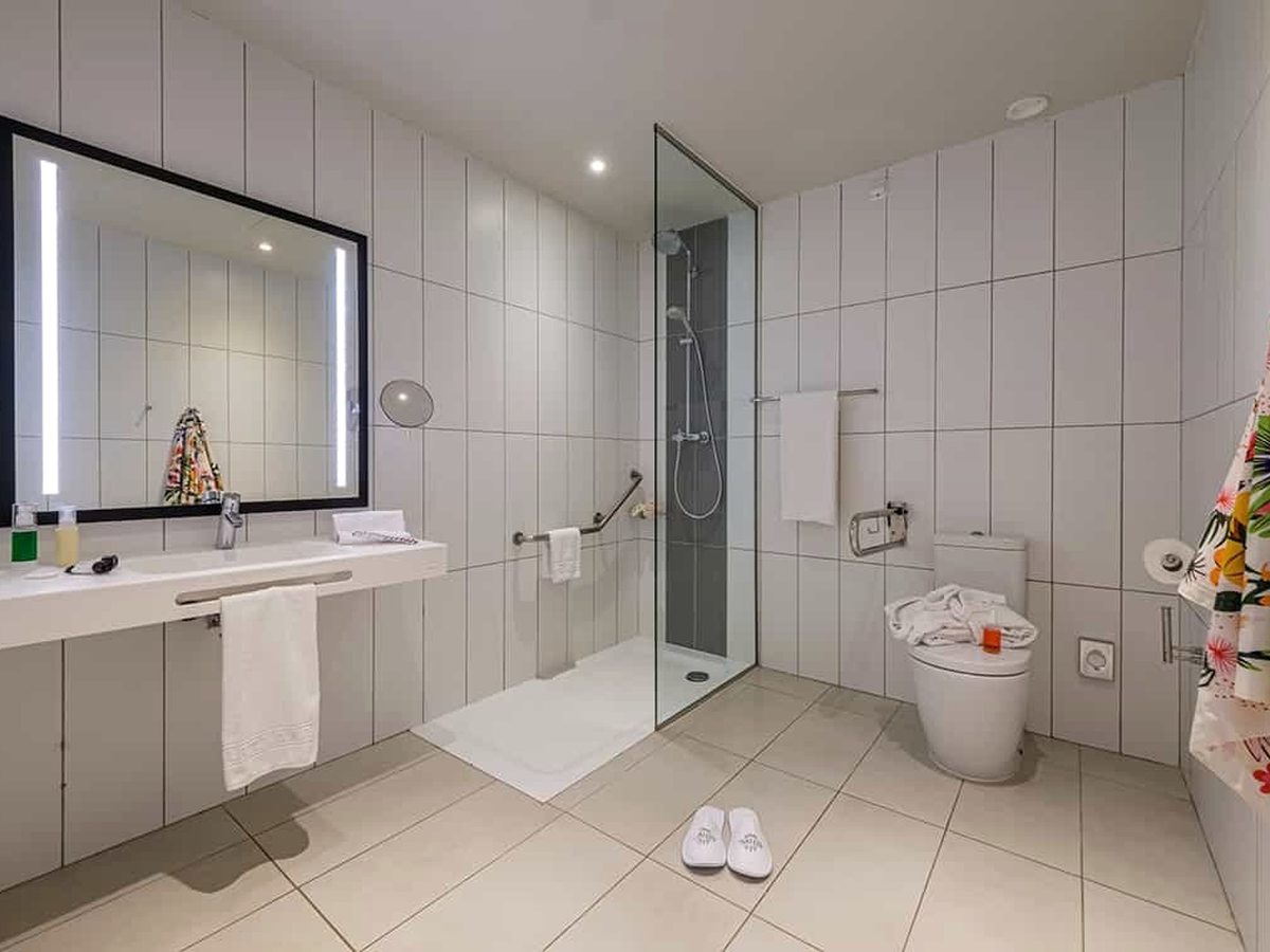 A white-tiled bathroom with a large lit mirror, a wide vanity, a glass walk-in shower and a grab rail.