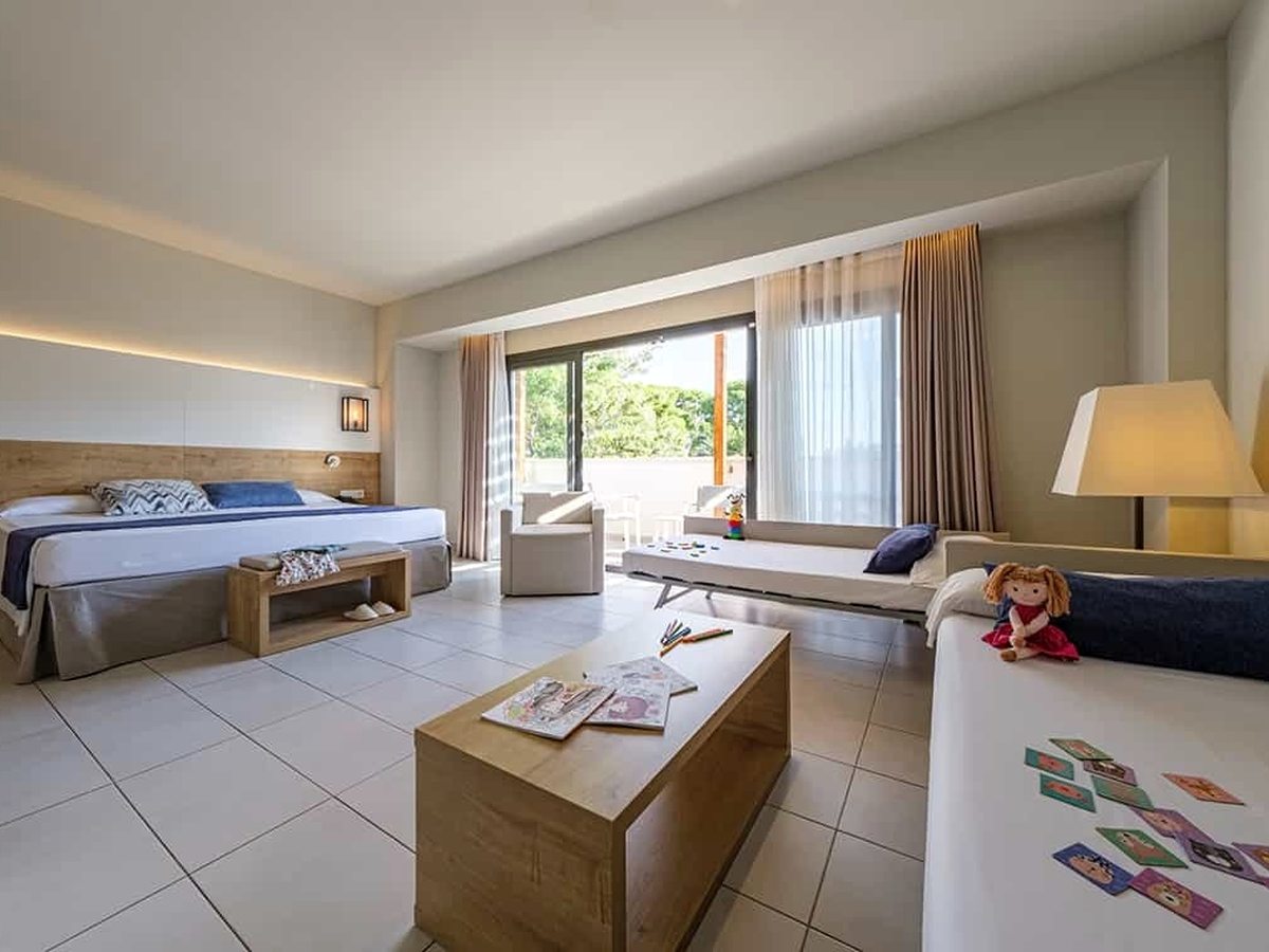 A spacious open-plan family room with a double bed, a sofa bed and a low table, children's books, crayons and a rag doll laid out, with a balcony beyond.