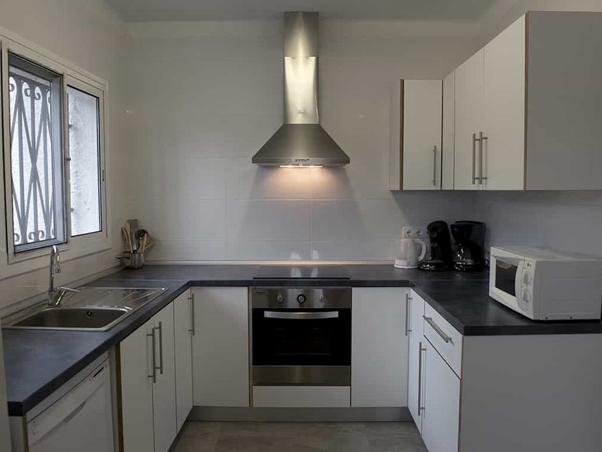 A compact fitted kitchen with white units, a dark worktop, an oven and hob, an extractor hood, a microwave, kettle and coffee machine, and a barred window.