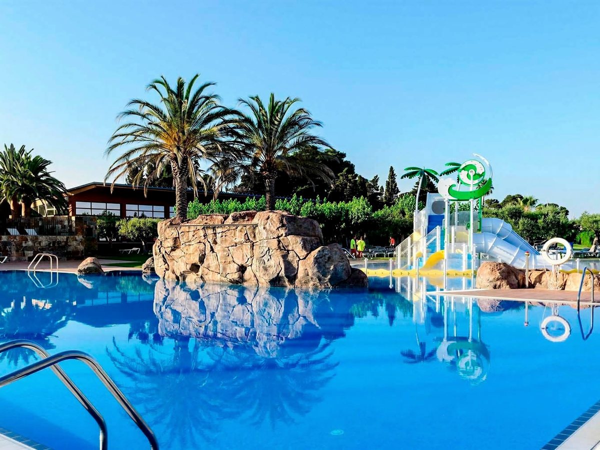 A large children's pool with a rocky central feature and a play structure with small slides and water toys, ringed by tall palms.