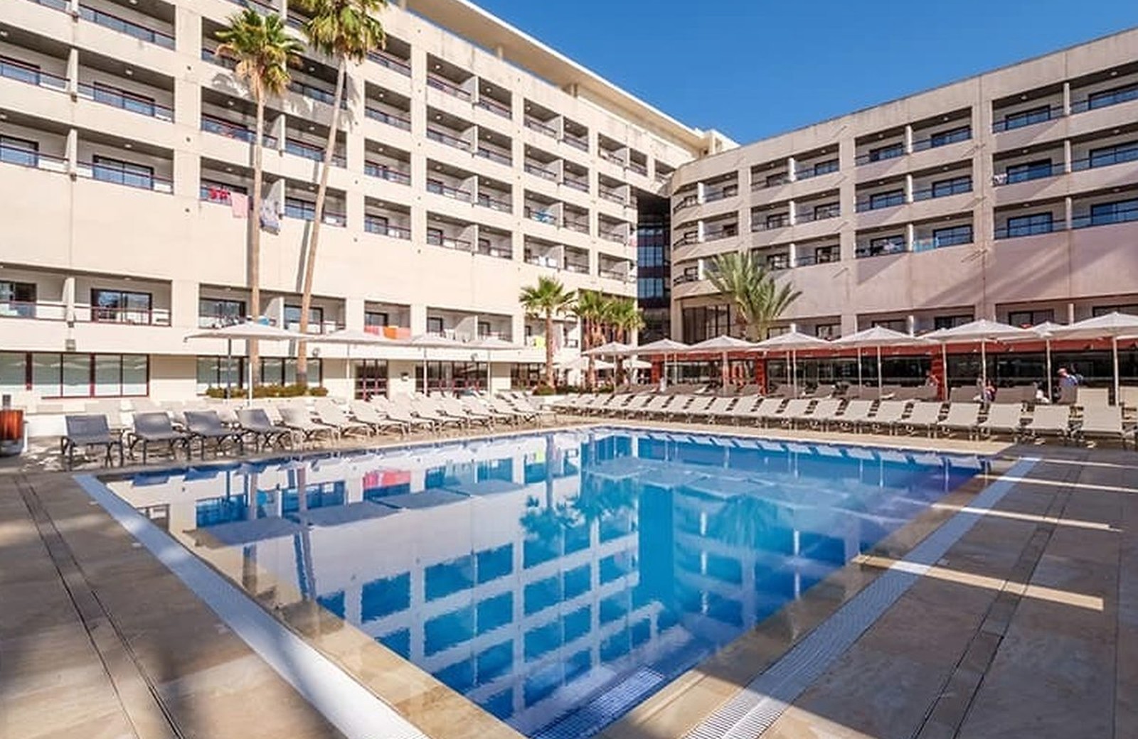 The main pool at Estival ElDorado below the tiers of hotel balconies, edged with sun loungers and umbrellas under a clear blue sky.
