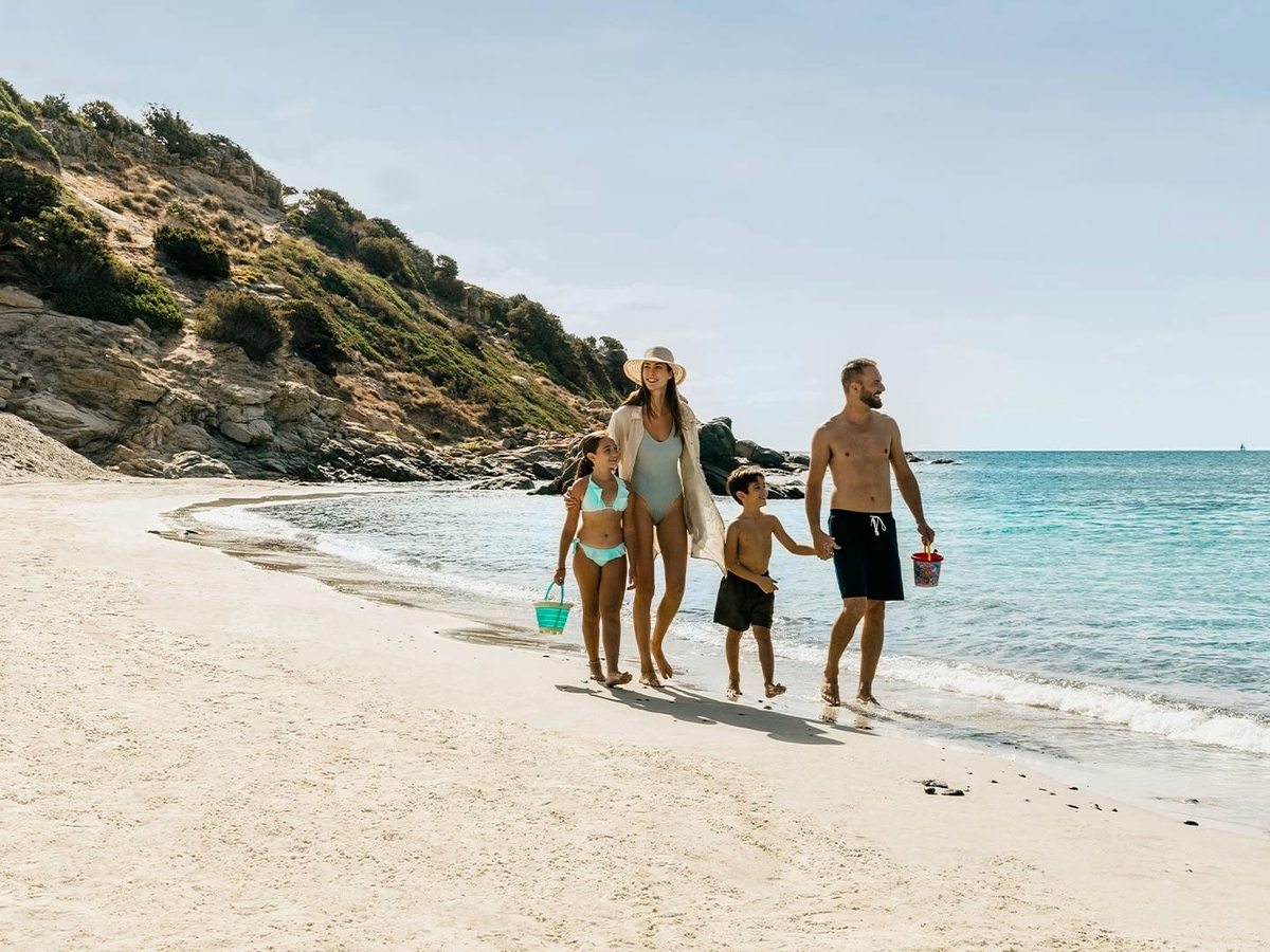 A family walking along a curving white-sand beach carrying buckets, clear shallow water to one side and green hills behind.