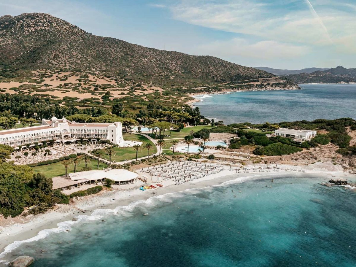An aerial view of the resort's crescent beach lined with parasols, backed by pine woods and hills, turquoise water in the foreground.