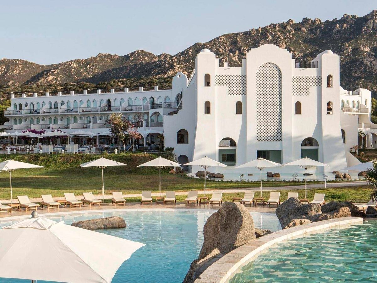 The white Moorish-styled main hotel building with a tall latticework tower, seen across an outdoor pool with parasols and loungers, hills behind.