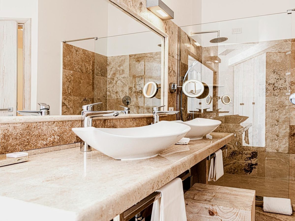 A twin-basin vanity on a travertine counter below a large mirror, with folded towels and travertine-tiled walls.