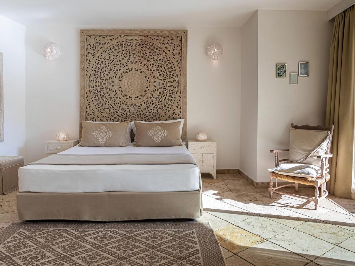 A double bed against a carved wooden latticework headboard, with matching bedside chairs and a patterned rug on a tiled floor.