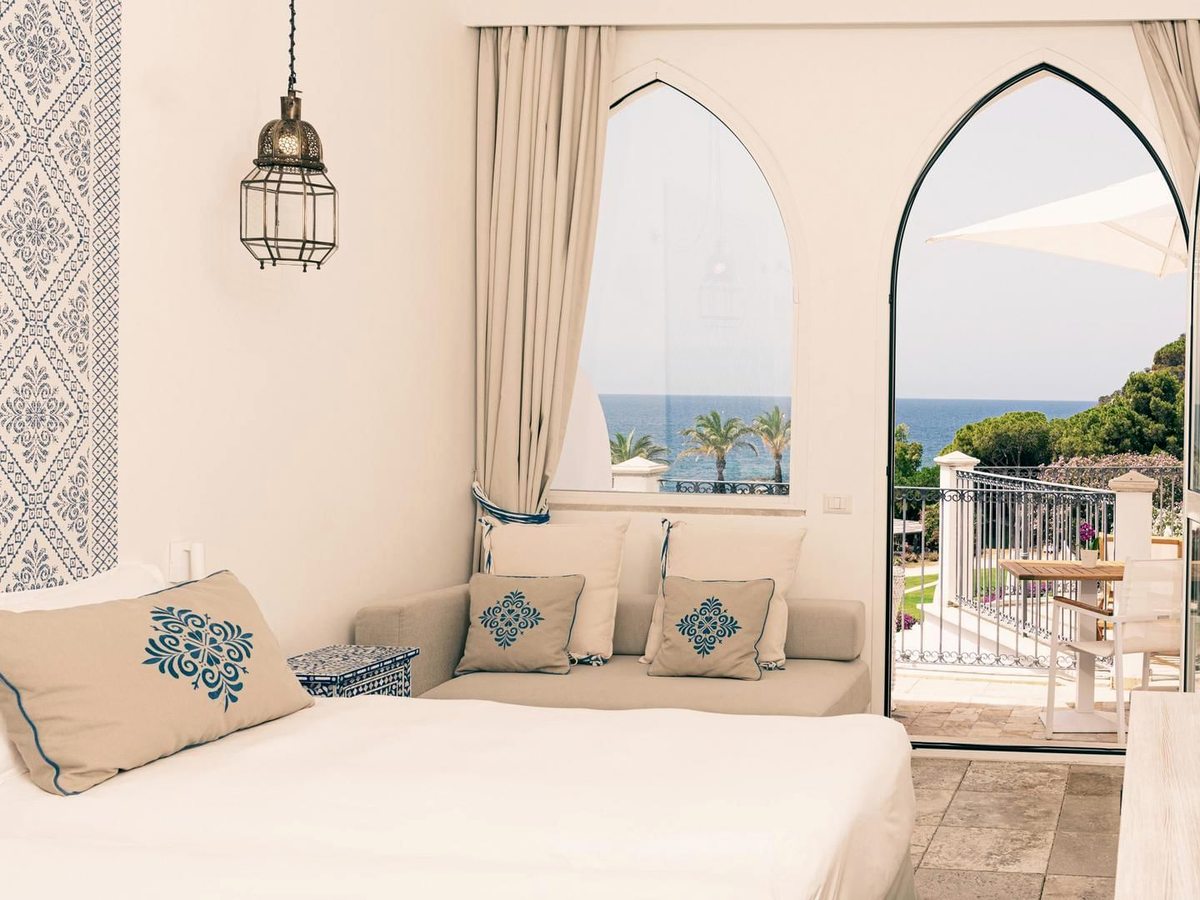 A cushioned corner seat beside a Moorish pointed-arch doorway opening onto a terrace with palms and the sea beyond.