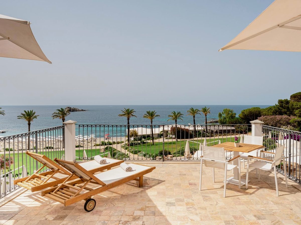 Wooden sun loungers and small tables on a travertine terrace, with lawns, palms and the sea beyond.