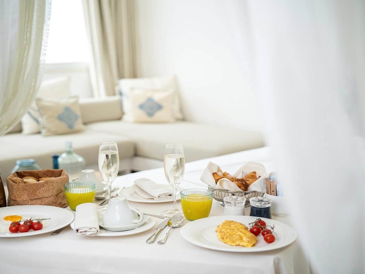 A breakfast plate of scrambled egg and tomatoes with a glass of orange juice, pastries and cherry tomatoes on a white-linen table.