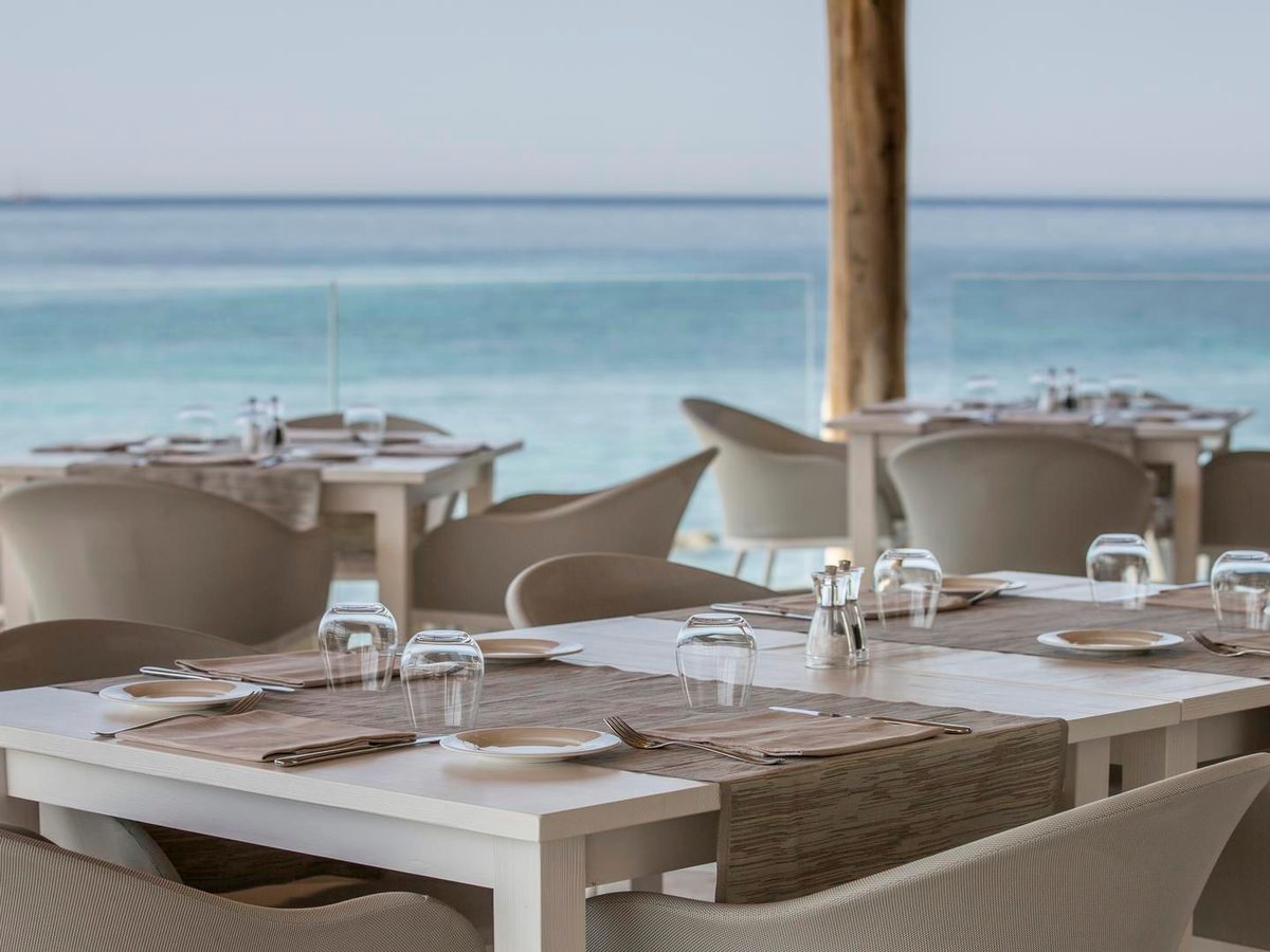 Set tables with wine glasses and folded napkins under a timber canopy, the turquoise sea directly beyond a glass balustrade.