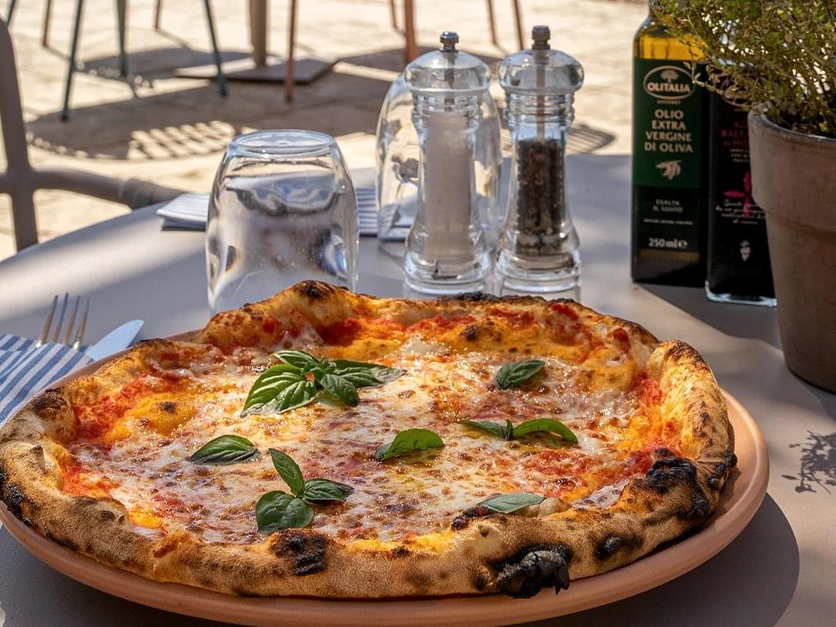 A tomato-and-basil pizza on a wooden board beside cutlery and cruets on a table set outdoors.