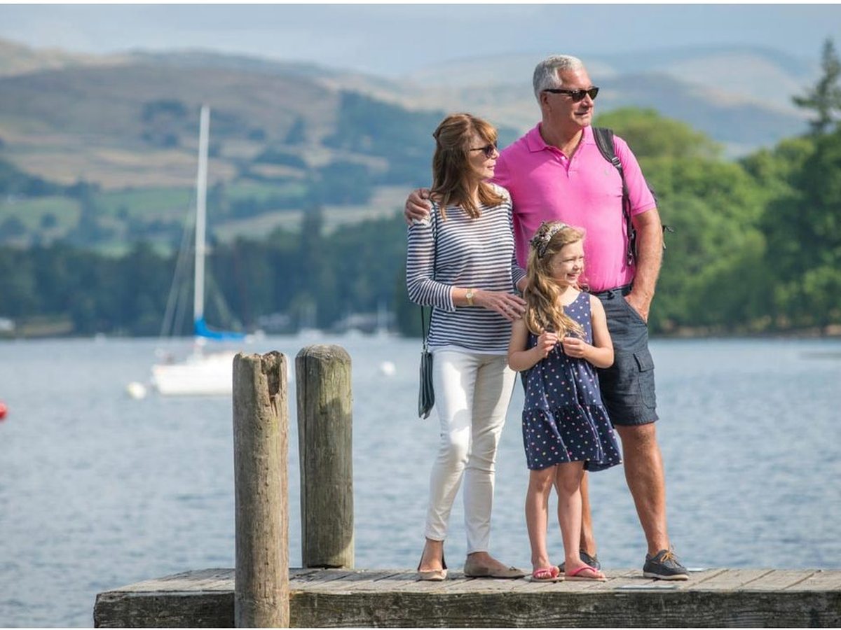 A family of three standing at the end of a wooden jetty, looking out across Lake Windermere to green fells, a moored yacht on the water behind them.