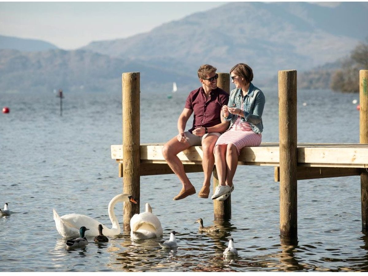 Two people sitting on the edge of a wooden jetty over the lake, with swans, ducks and gulls gathered on the water below and fells rising beyond.