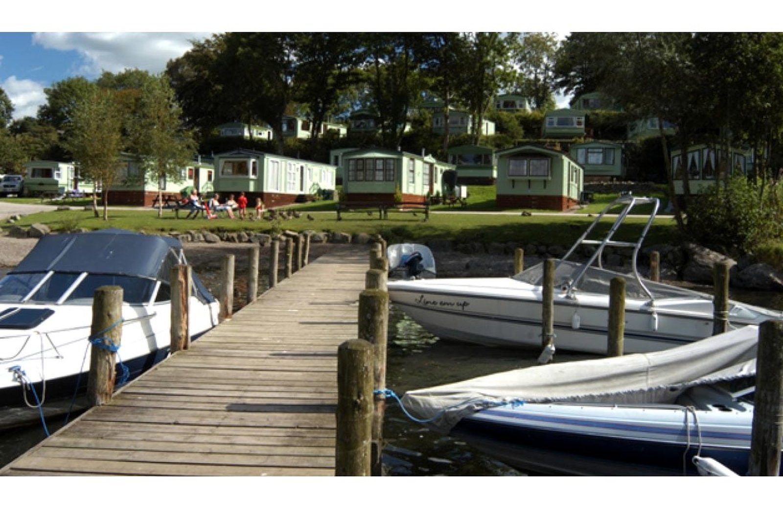 Fallbarrow Holiday Park: caravans and lodges set on a wooded, terraced estate running down to a marina on the eastern shore of Lake Windermere, with boats moored and wooded fells beyond.