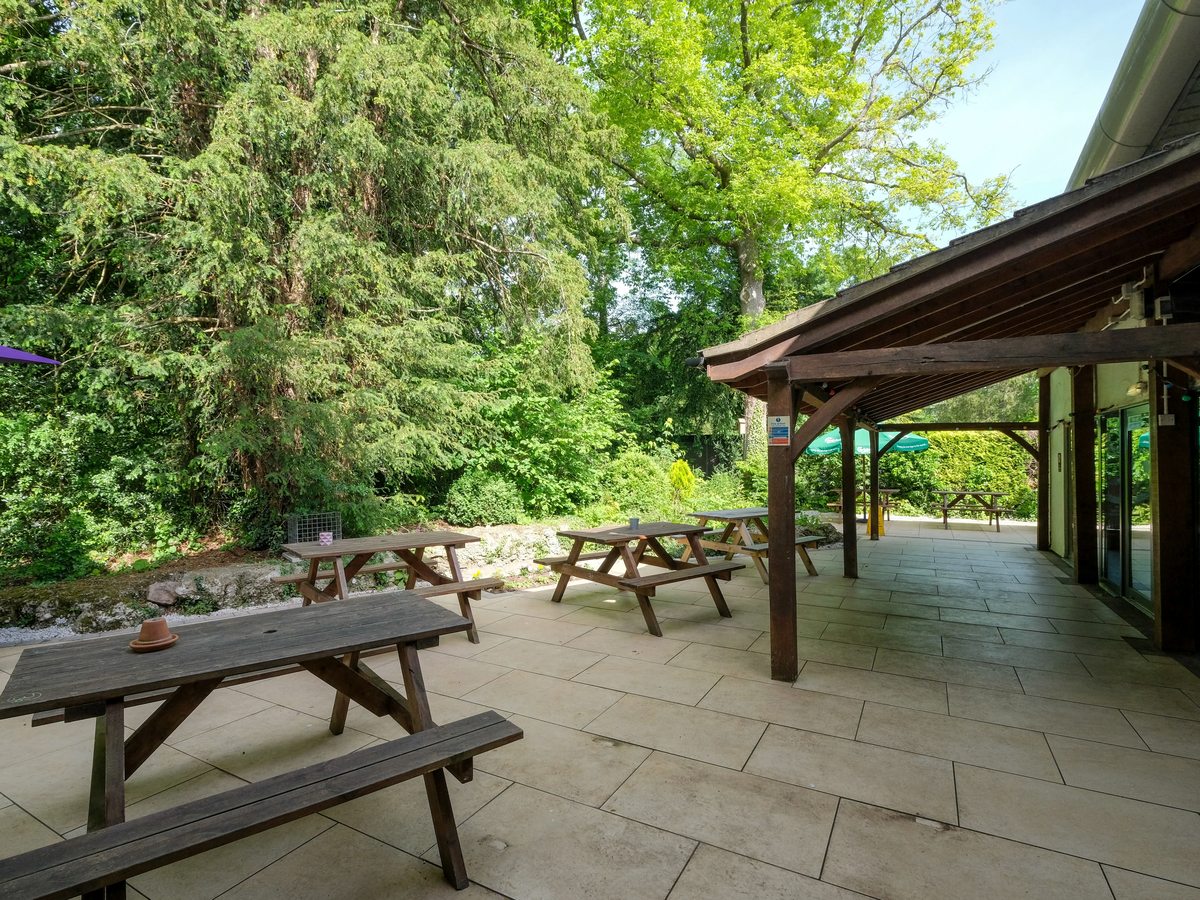 A covered timber terrace with picnic benches on a paved patio beside trees and a limestone rockery.