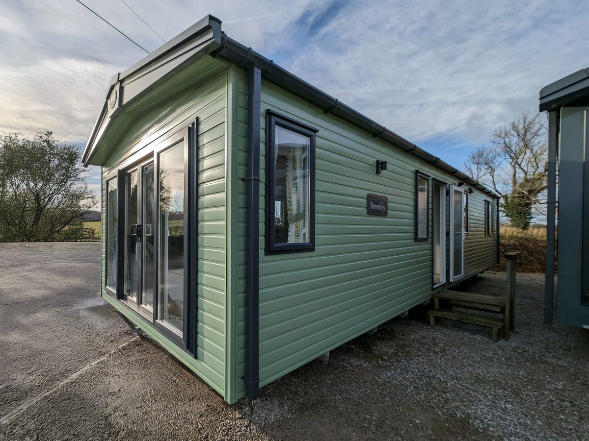 A pale green static caravan with large front windows and a decked veranda on a gravel pitch.