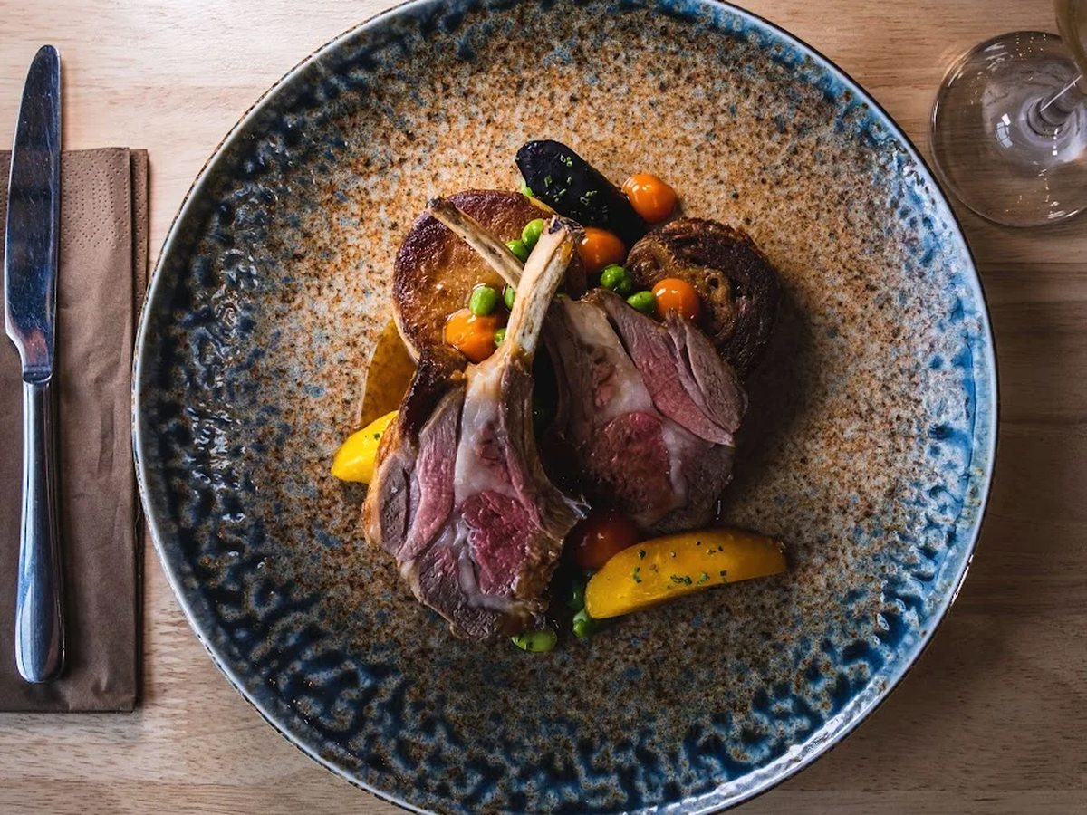 A plated rack of lamb with peas, roasted vegetables and a wine glass on a wooden table.