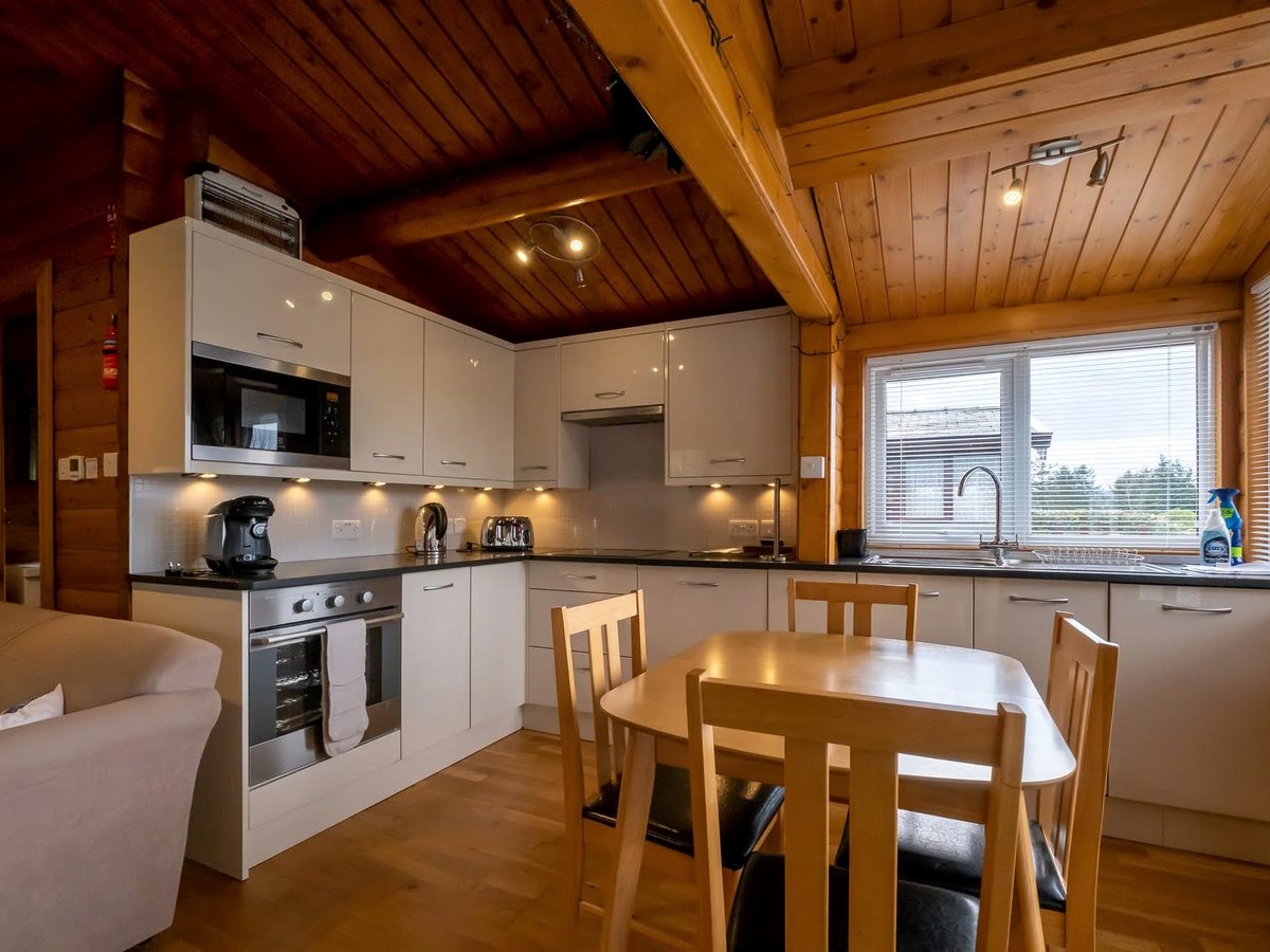 A fitted lodge kitchen with white units, oak worktops, an oven and a small dining table under a vaulted timber ceiling.