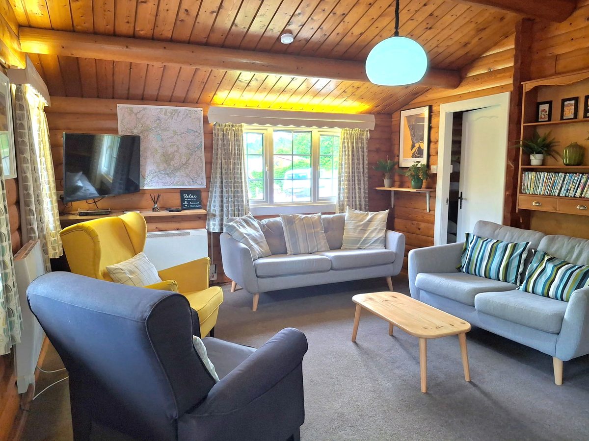 A wood-clad lodge lounge with a vaulted timber ceiling, grey and yellow armchairs and a Lake District map on the wall.