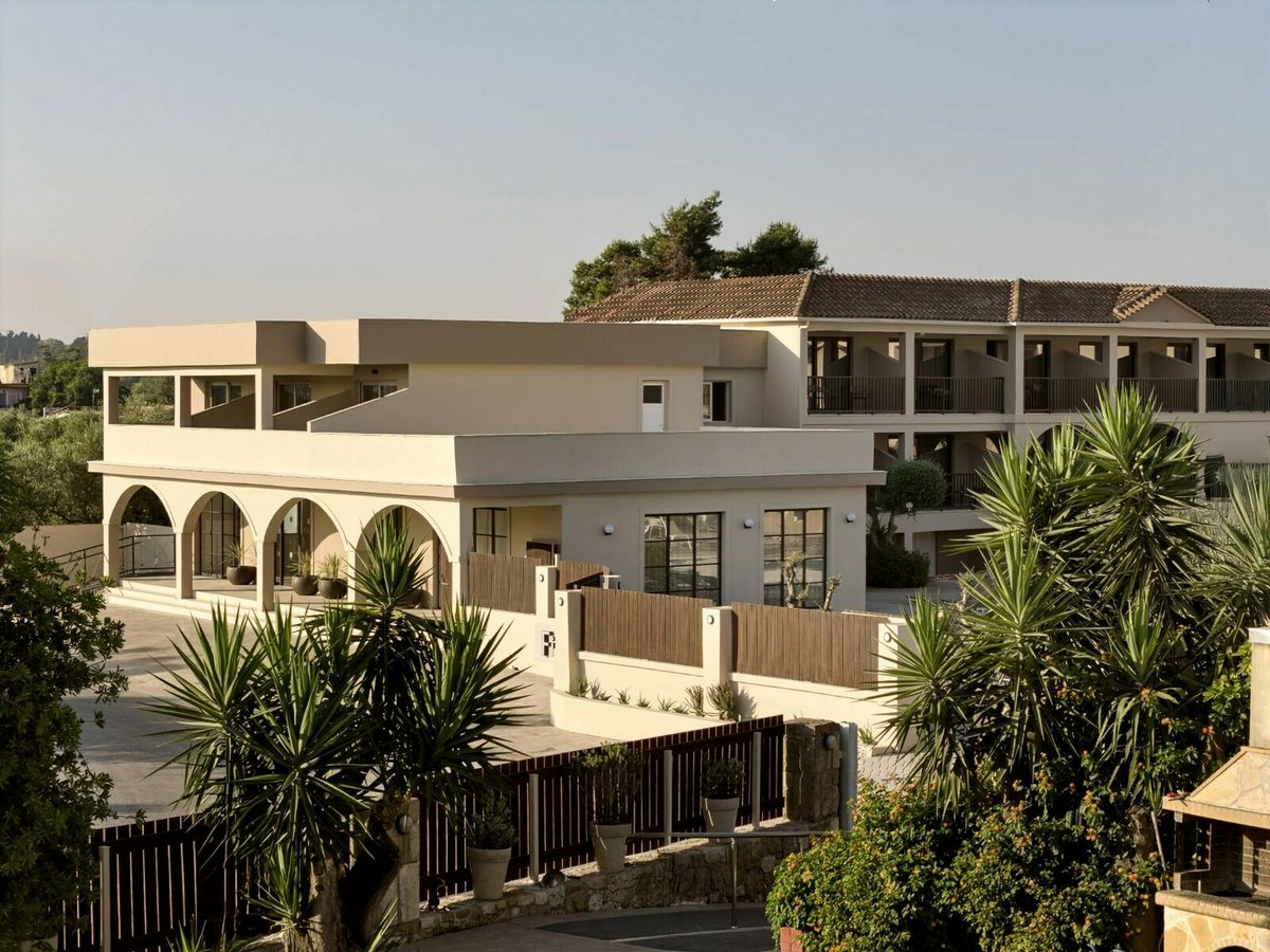 A cream-rendered low-rise hotel block with an arched ground-floor colonnade and a balconied upper storey, palms and bamboo screening in front.