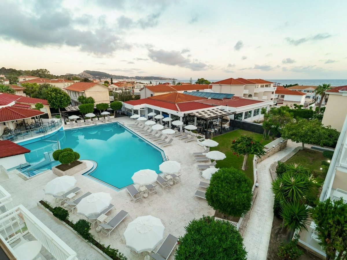 Aerial view of Filoxenia Hotel with a rectangular pool ringed by white parasols and loungers, gardens, red-roofed low-rise buildings and the sea in the distance.