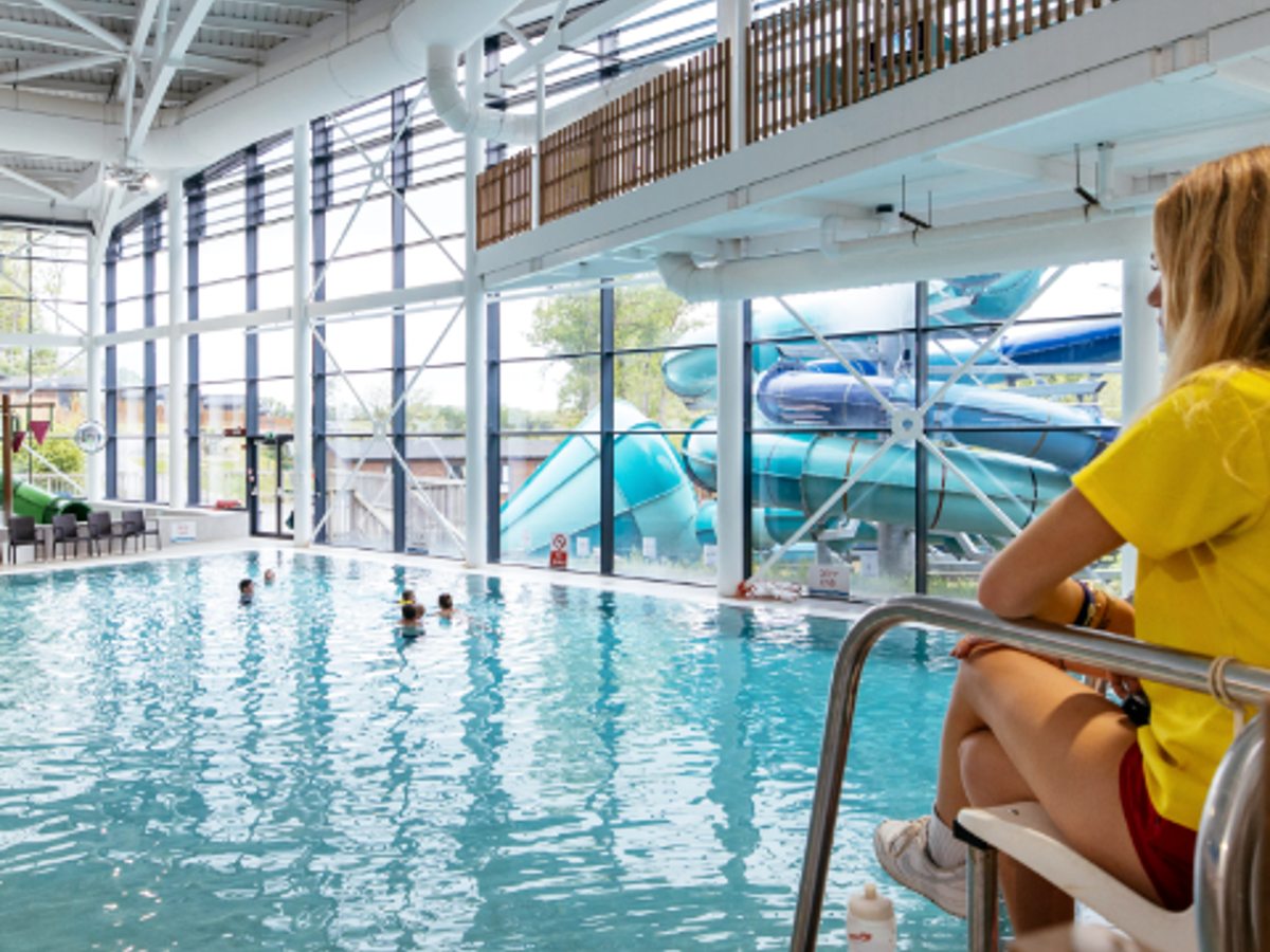 A high-ceilinged indoor pool with a glass wall, a lifeguard on a raised chair and blue flumes visible outside