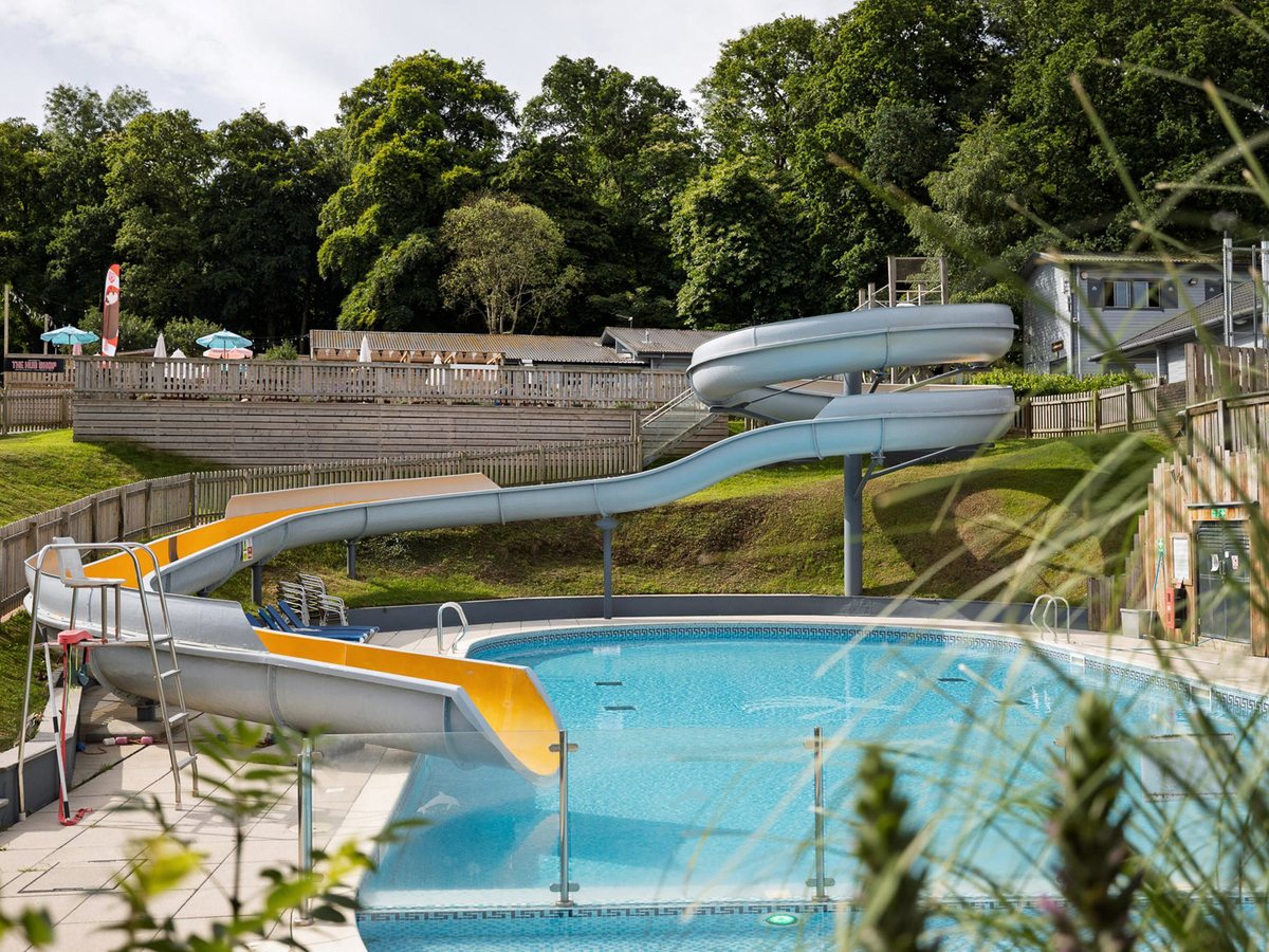 An outdoor pool with a grey and yellow flume slide, backed by tall woodland