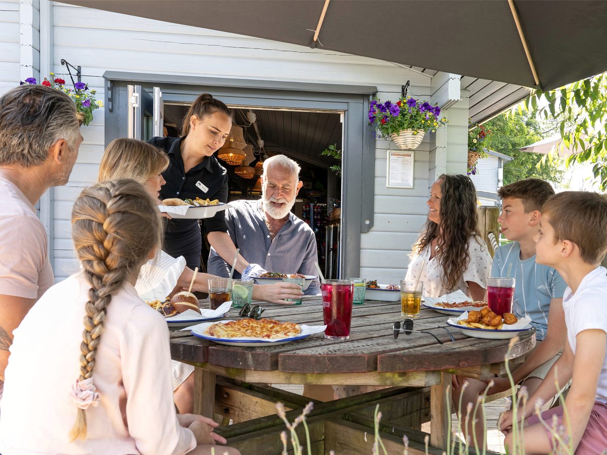 A family at a round wooden table under a parasol on a covered veranda, a member of staff bringing food