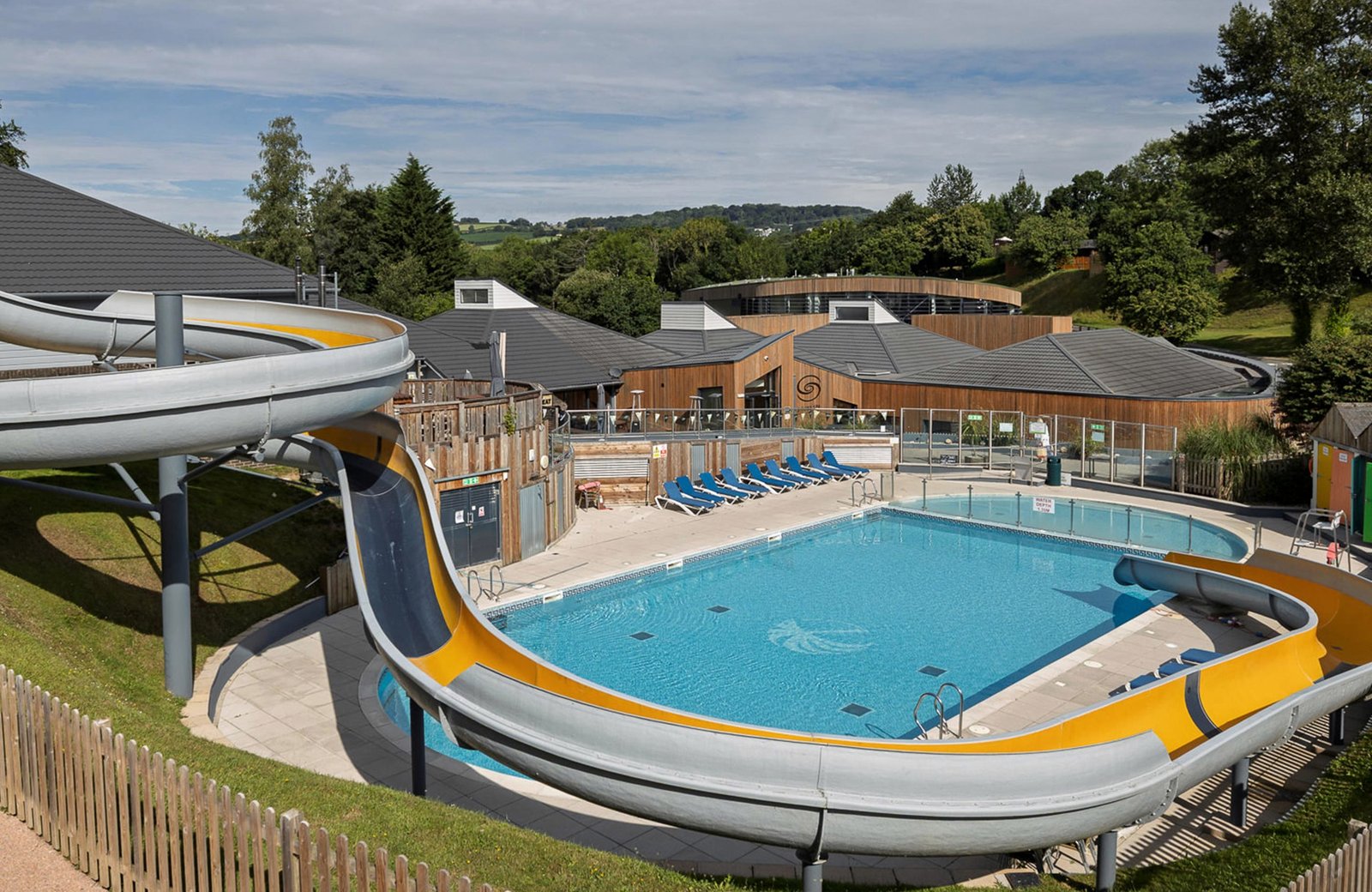 The outdoor pool at Finlake, with grey and yellow flume slides, sun loungers and the resort buildings set among wooded Devon hills.