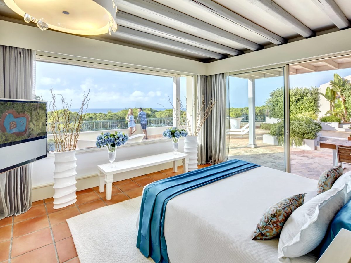A bright bedroom with a beamed ceiling and corner windows opening to a large terrace overlooking gardens and the sea