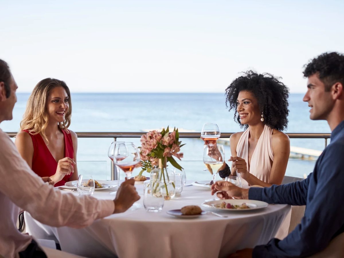 Four adults dining at a white-clothed table on a restaurant terrace with the sea behind them